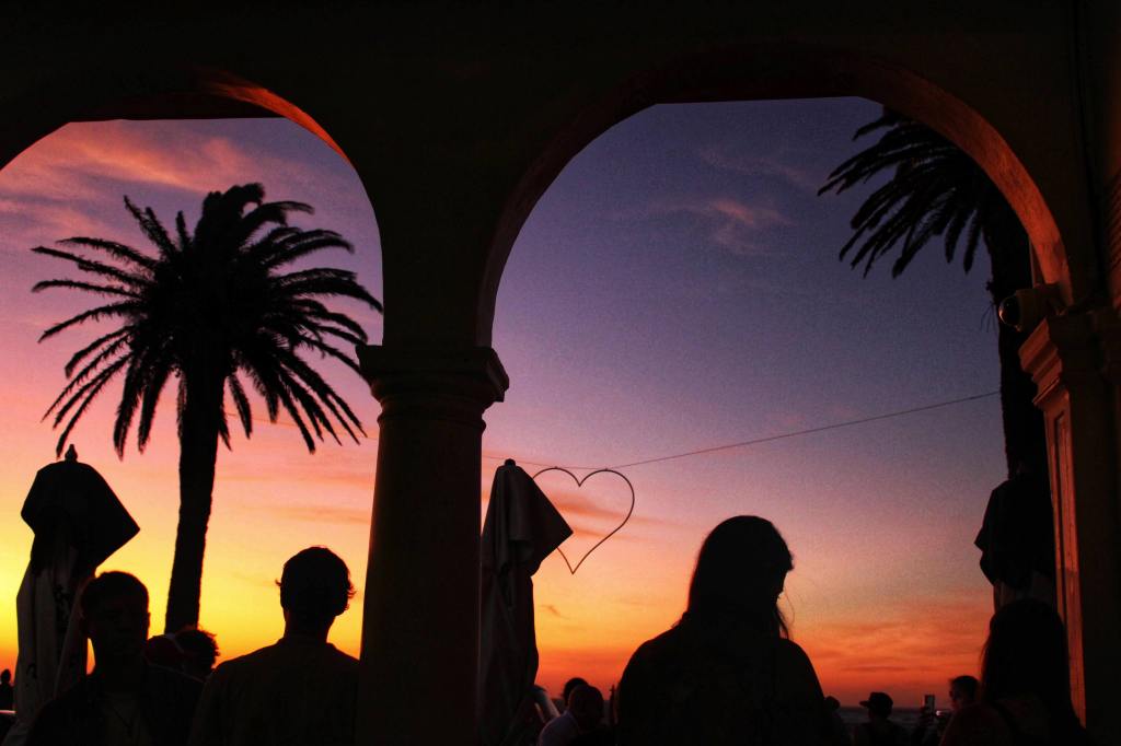 A colorful sunset in Camps Bay with silhouettes palm trees, a heart hanging in the sky and cafe goers.